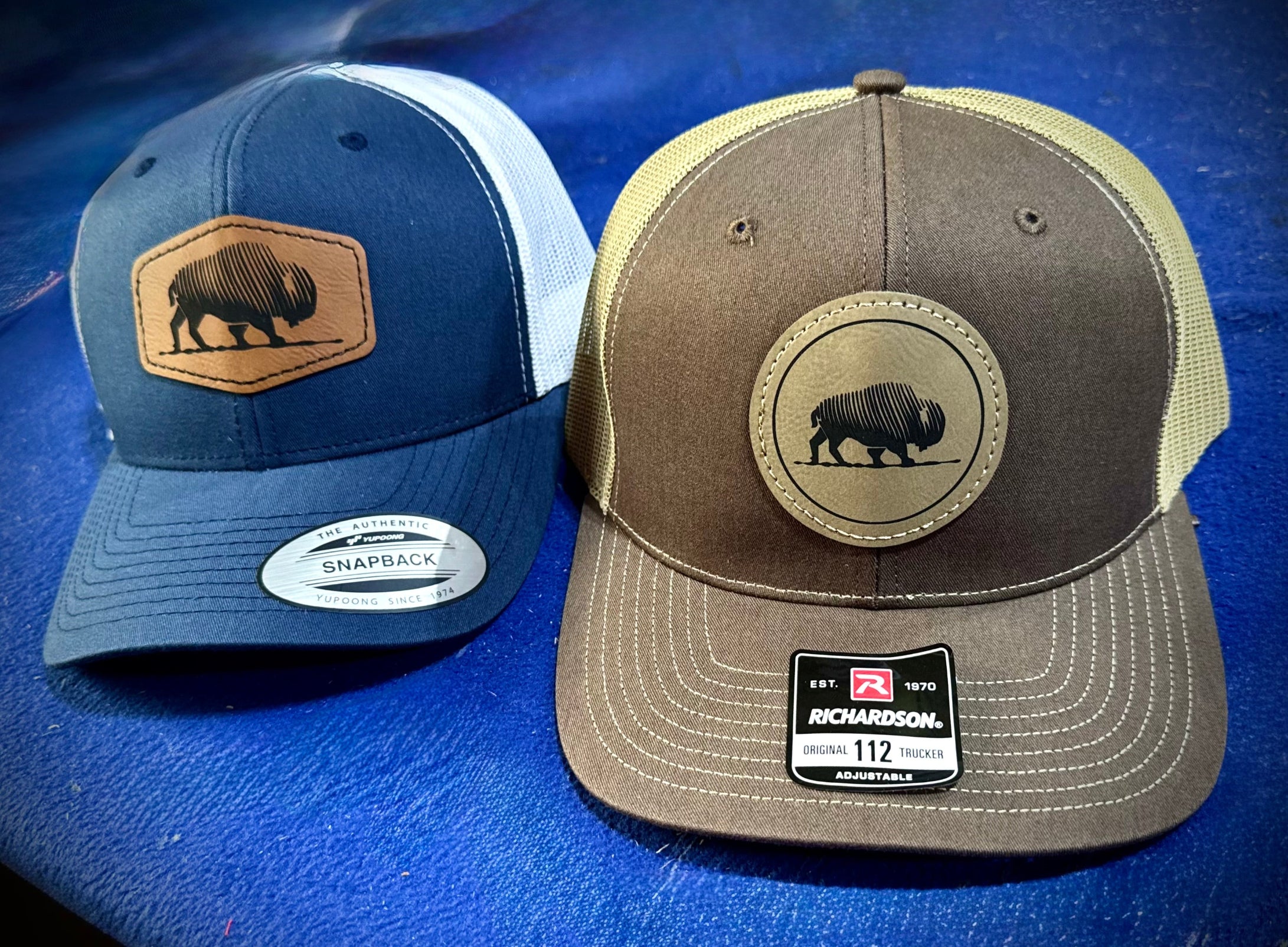 Two baseball caps with leather bison patches on a blue background