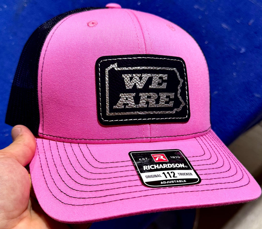 Pink cap with 'WE ARE' patch held against a blue background