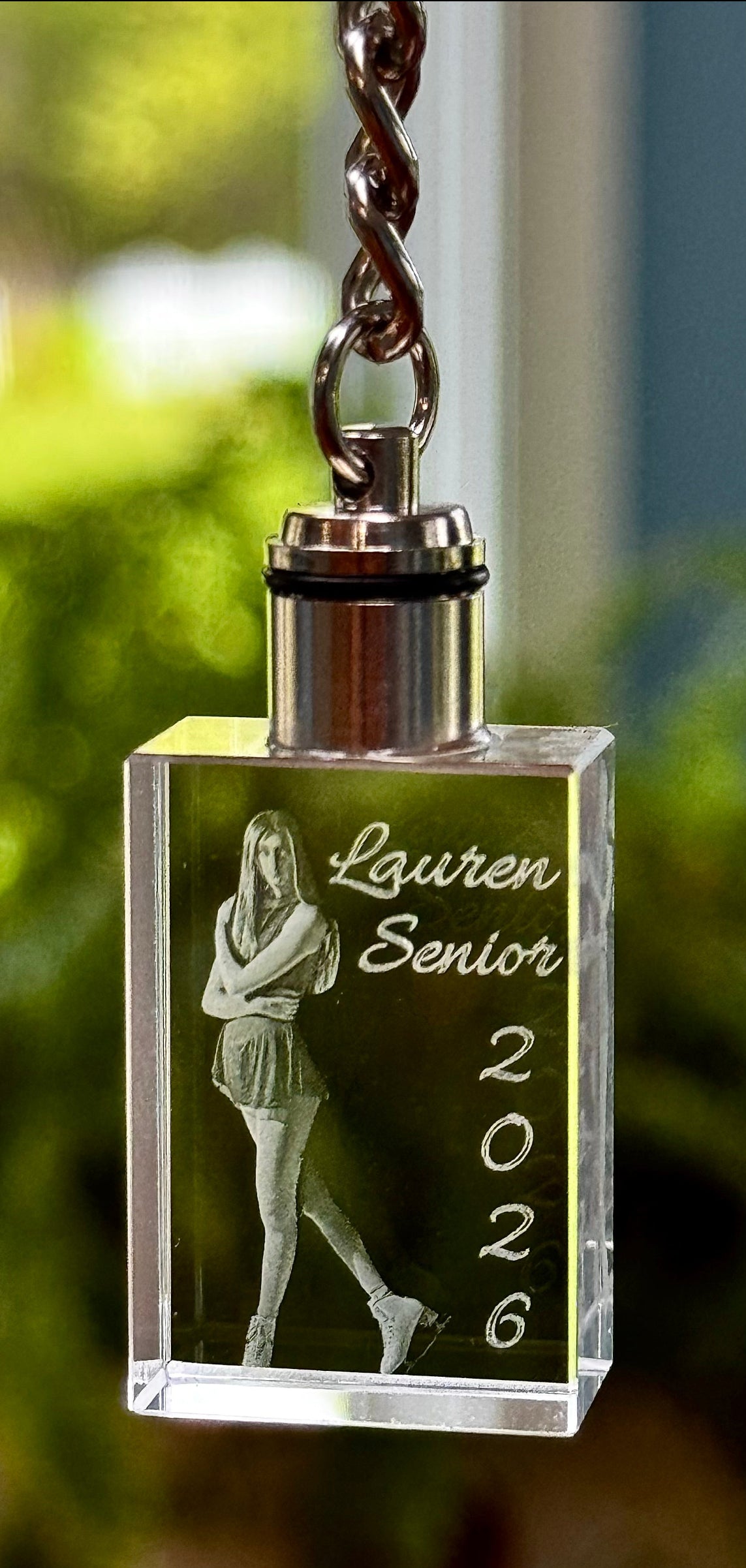 Crystal keychain with engraved photo and text 'Lauren Senior 2026' against a blurred green background.