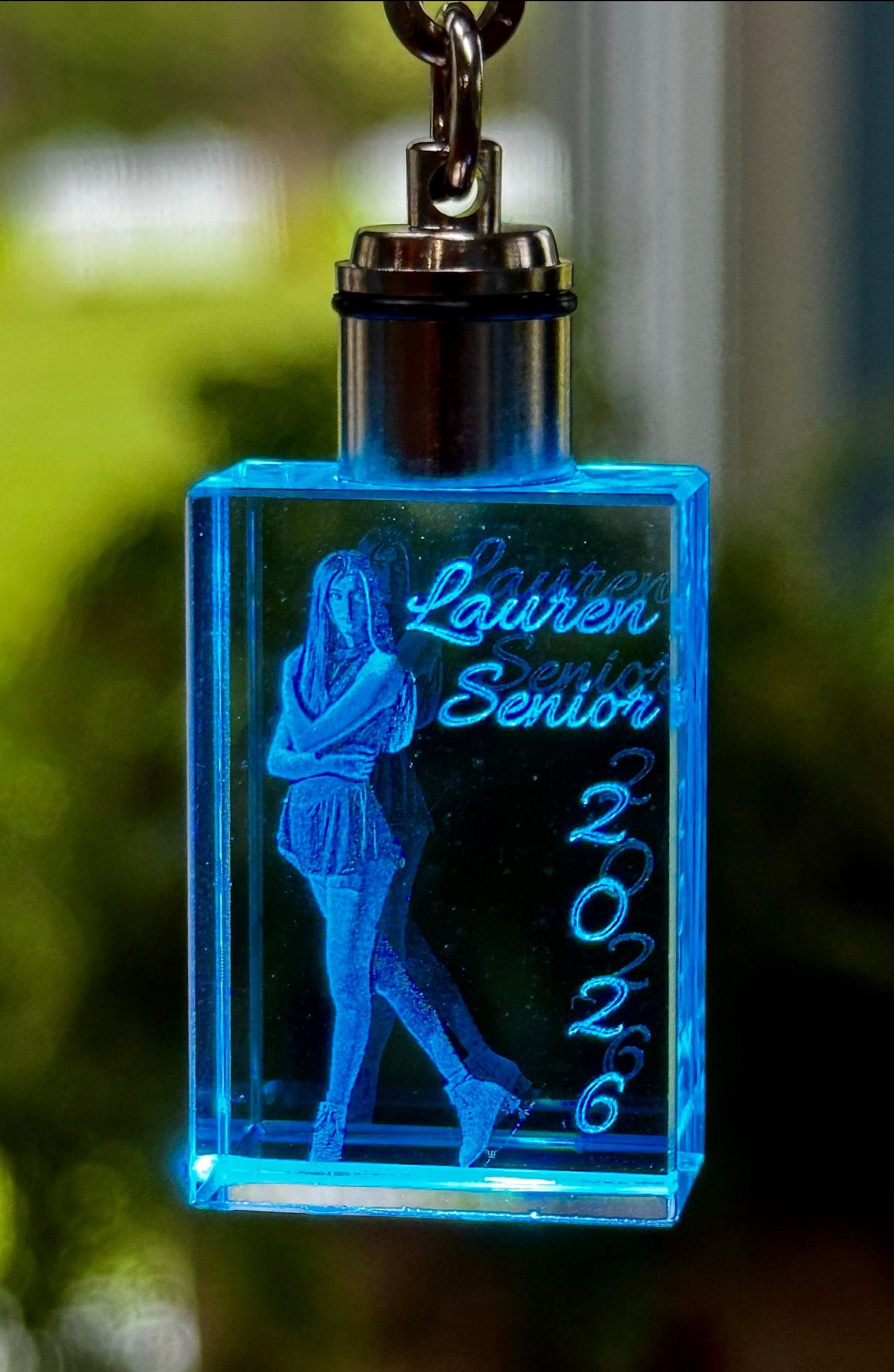 Blue LED keychain with engraved text and silhouette of a person, blurred green background
