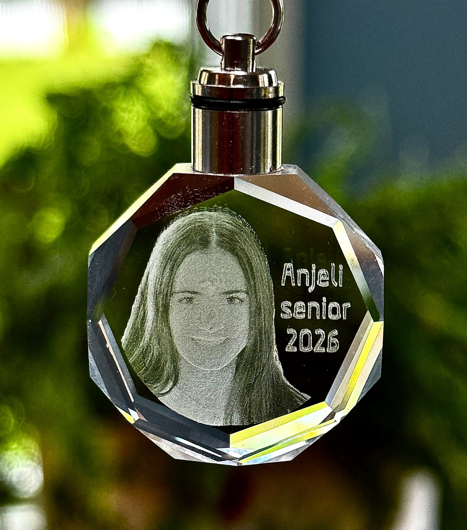 Crystal keychain with engraved photo and text 'Anjeli senior 2026' against a blurred green background